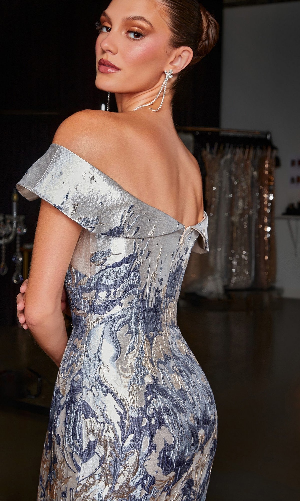 A woman indoors, glancing over her shoulder, wears the Ladivine CD0107 long formal dress in silver and blue with an off-the-shoulder neckline and subtle mermaid silhouette; blurred clothing racks and dresses fill the background.